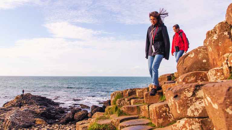 Two visitors walking along the Giant's Causeway's iconic basalt columns with the ocean in the background under a partly cloudy sky